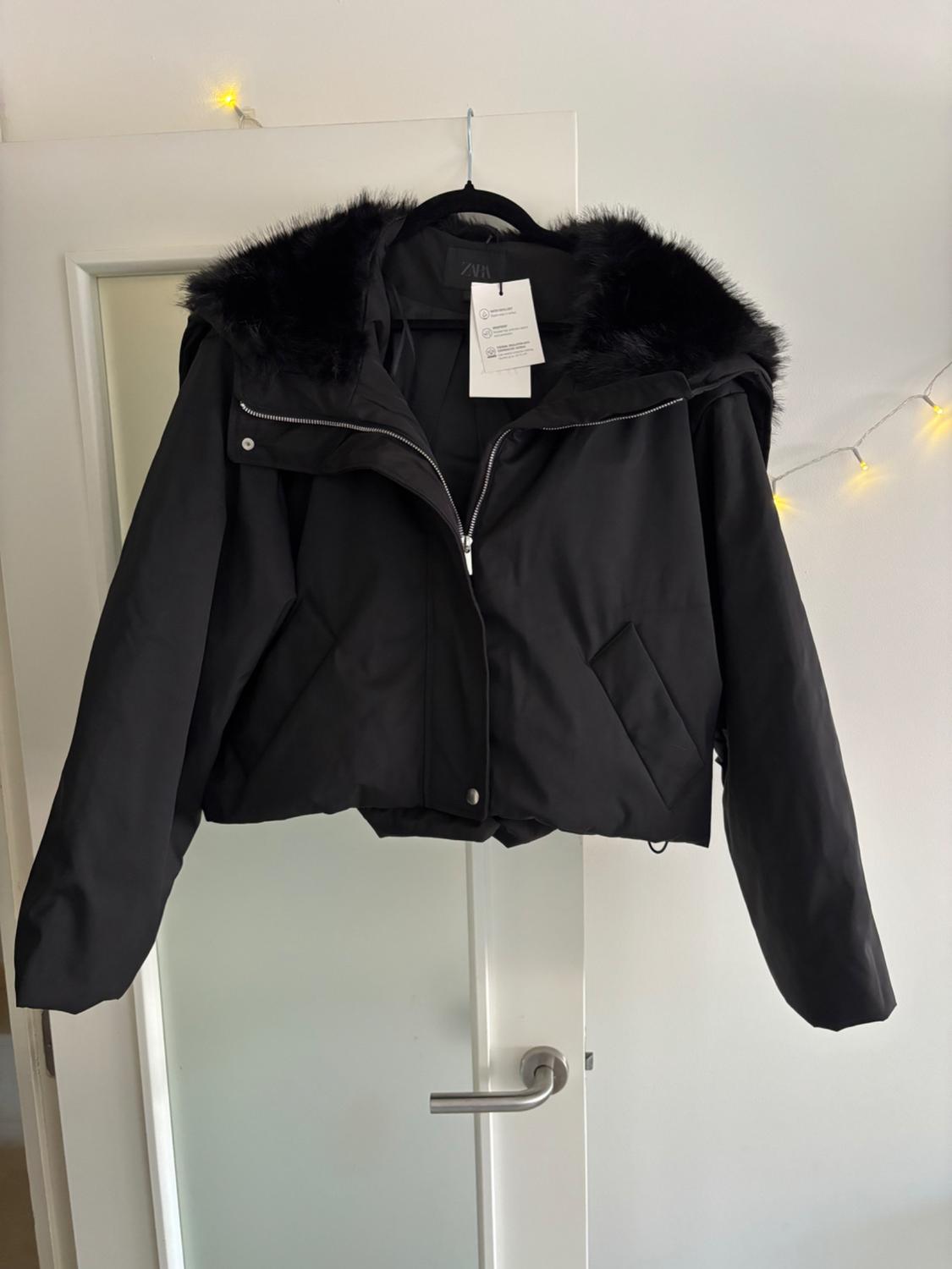Black jacket with a fur hood hanging on a white door. Decorative yellow lights in the background. Label visible.