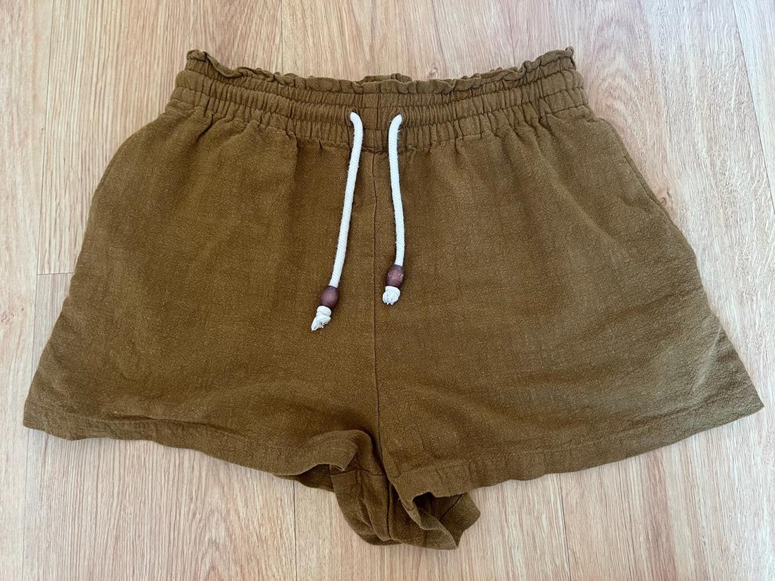 Relaxed-fit brown shorts with a gathered elasticated waist and a white drawstring with wooden beads, set against a light wood background.