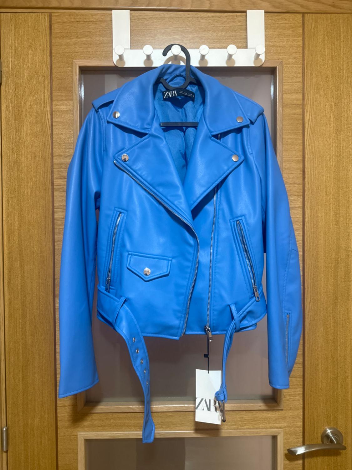 Blue jacket hanging on a wooden door. Silver zip and button details. Visible label with the text "ZARA".
