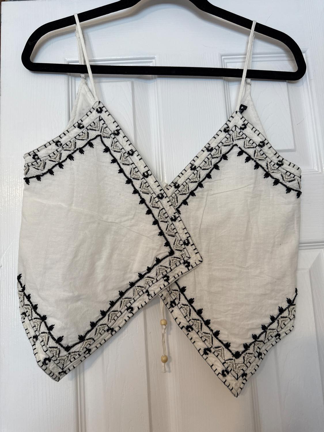 White top with spaghetti straps and black embroidery on the trim, hanging on a black hanger over a white door.