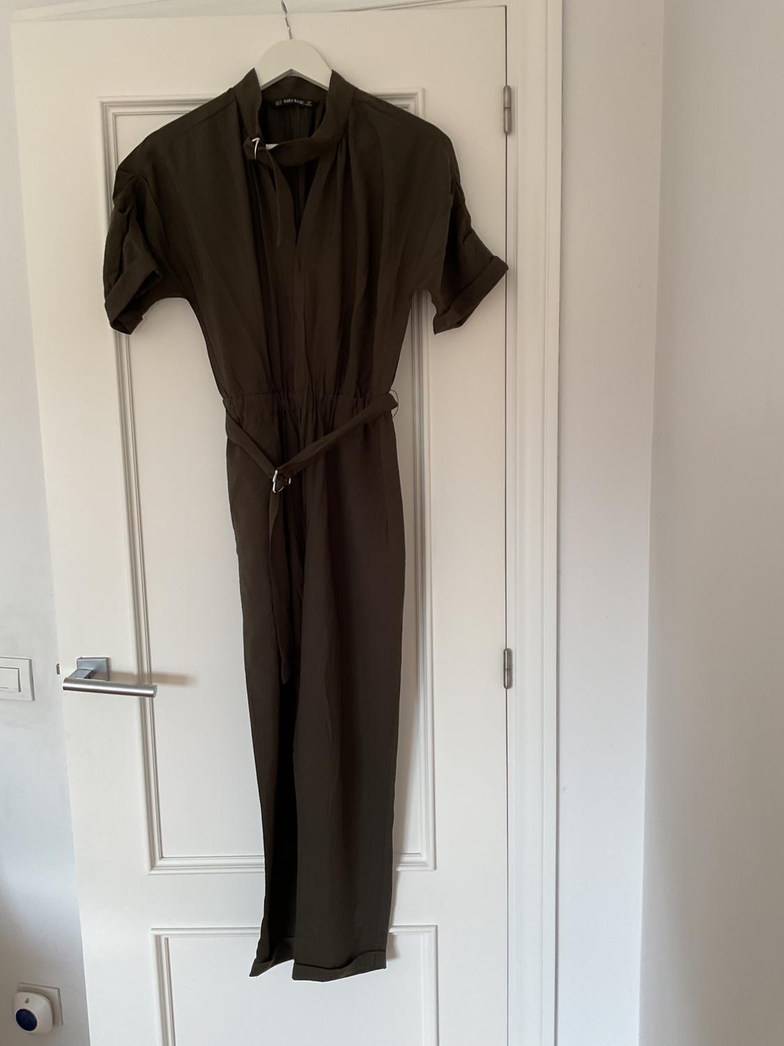 Olive green jumpsuit hanging on a white door, with a matching belt and short sleeves. Minimalist and simple setting.