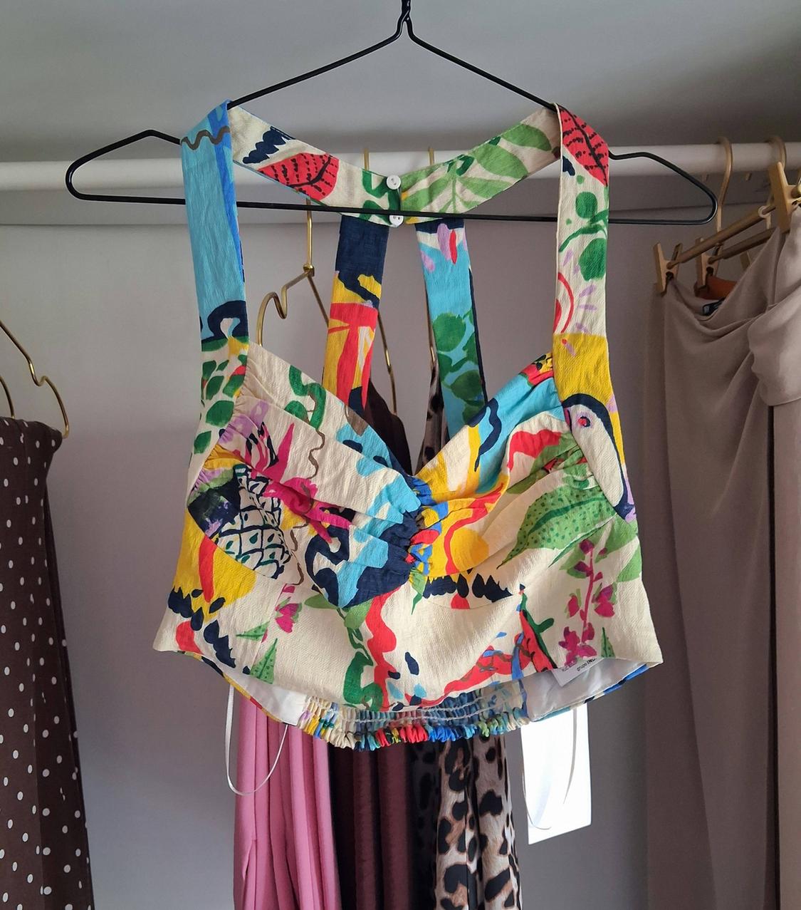 Multicoloured cropped camisole top, hanging on a black hanger on a clothes rail with other garments in the background. Relaxed atmosphere.