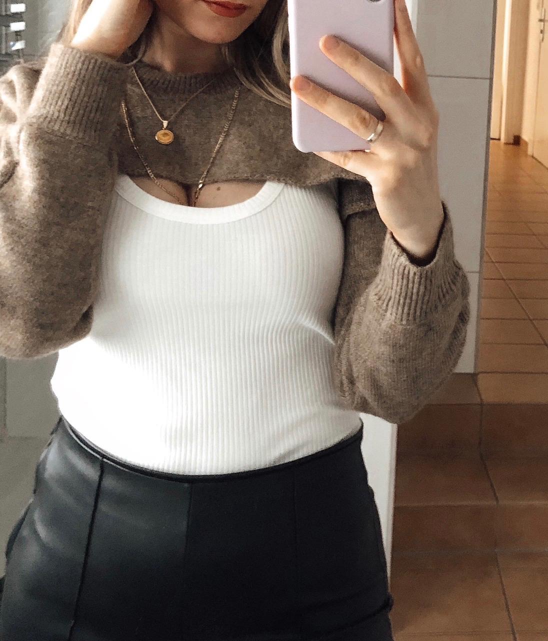 Woman taking selfie in mirror, wearing brown jumper, white ribbed top, black skirt, and gold necklaces. Pink phone in hand.
