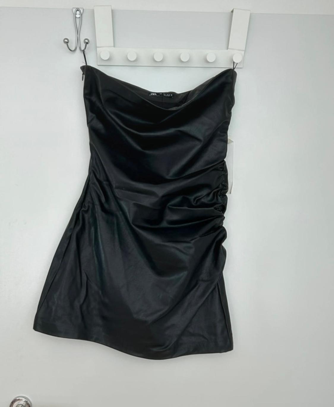 Black strapless dress hanging on a white clothes rail against a white wall. Shiny fabric with pleats at the side.