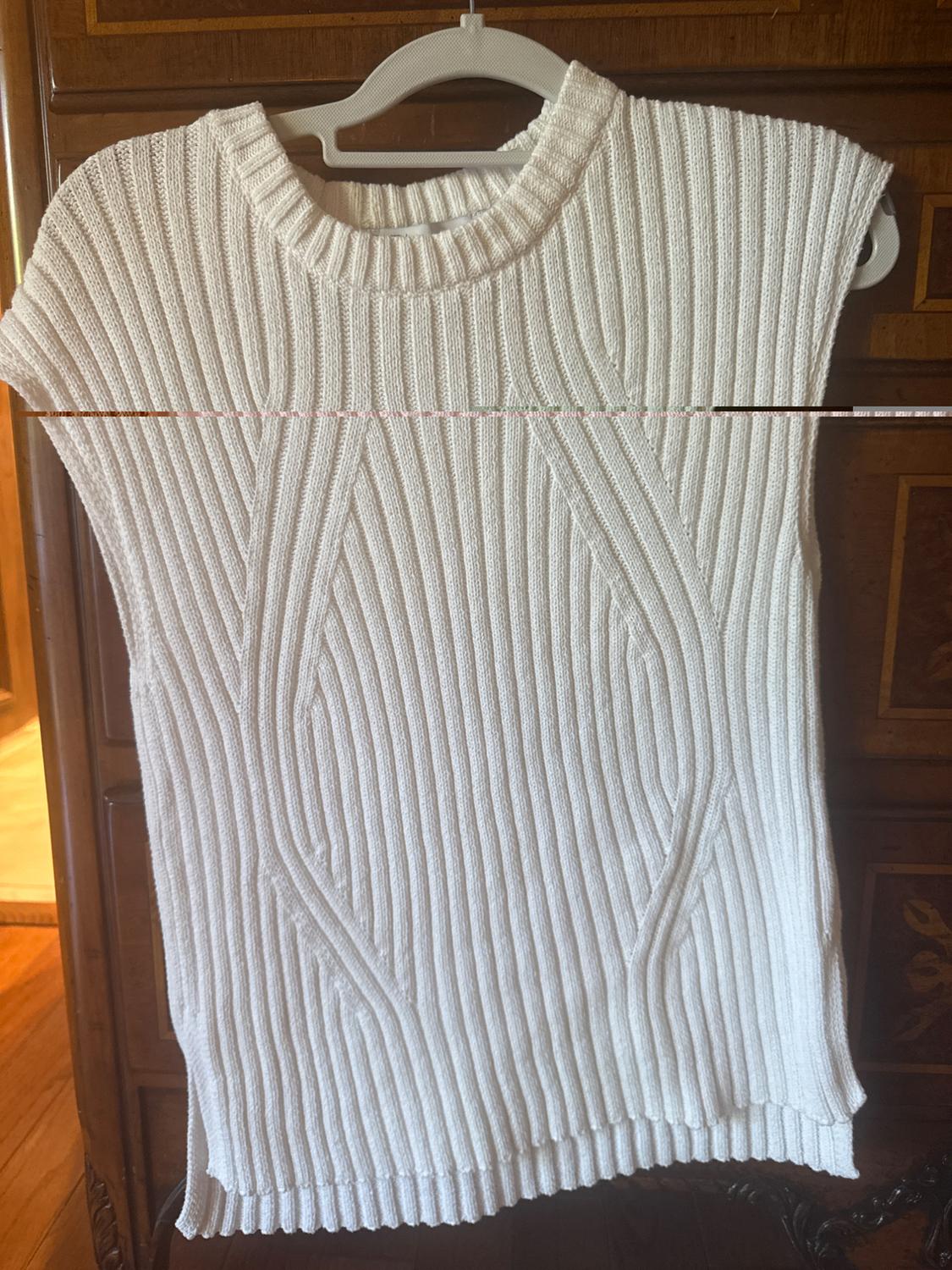 White ribbed knitted vest hanging on a white hanger, against a dark wooden background.