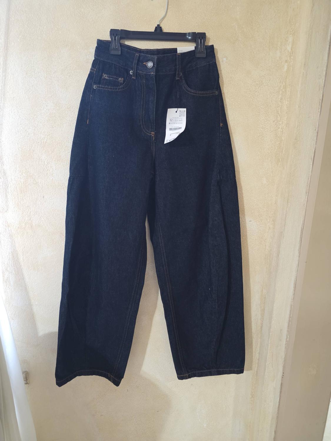 Dark blue wide-leg jeans hanging on a black hanger against a light beige wall.
The stitching and white label stand out.