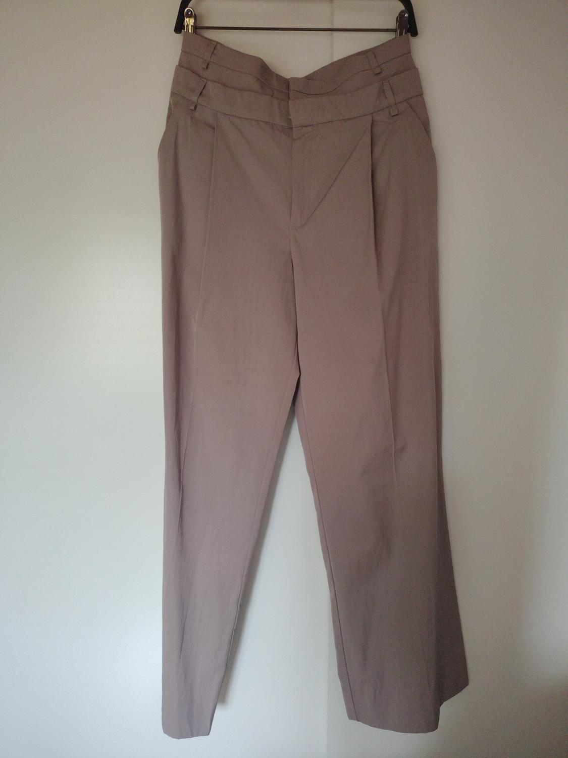 Beige trousers hanging on a hanger against a white background. High-waisted design with front pleats and button fastening.