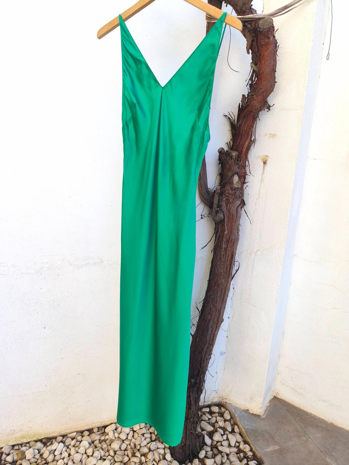 Green slip dress hanging on a wooden hanger, next to a white wall and a dry branch. Stone floor.
