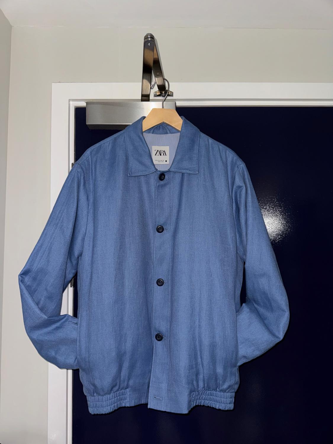 Blue button-up jacket hanging on a wooden hanger. Visible label with the text "ZARA". Dark background and white door.