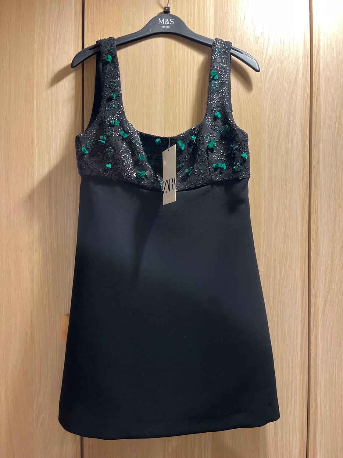Black dress with green sequin-embellished straps, hanging on an M&S hanger against a light wooden background.