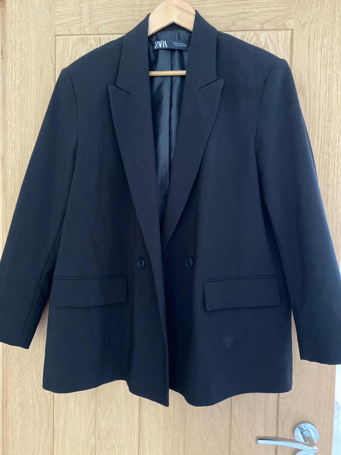 Black jacket hanging on a wooden hanger over a light wooden door. Visible label with the text "ZARA".