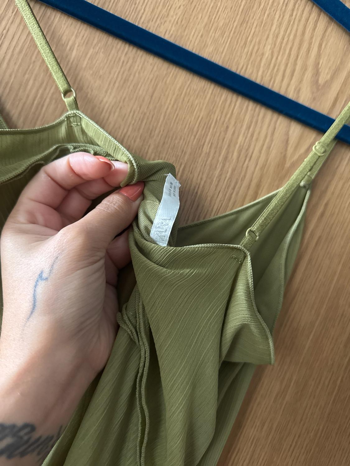 Hand holding the ZARA label of an olive green pleated strap dress on a blue hanger against a wooden background.