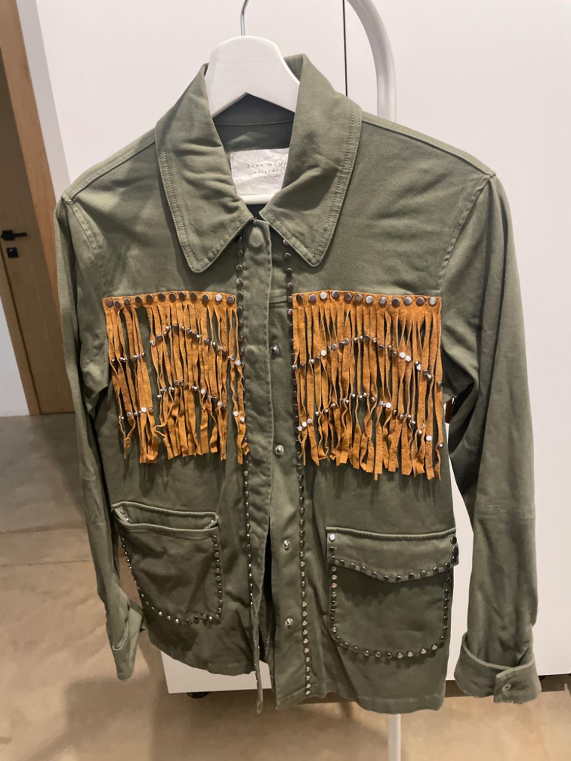 Green jacket with brown fringes and studs on the pockets and chest, hanging on a white hanger against a neutral background.