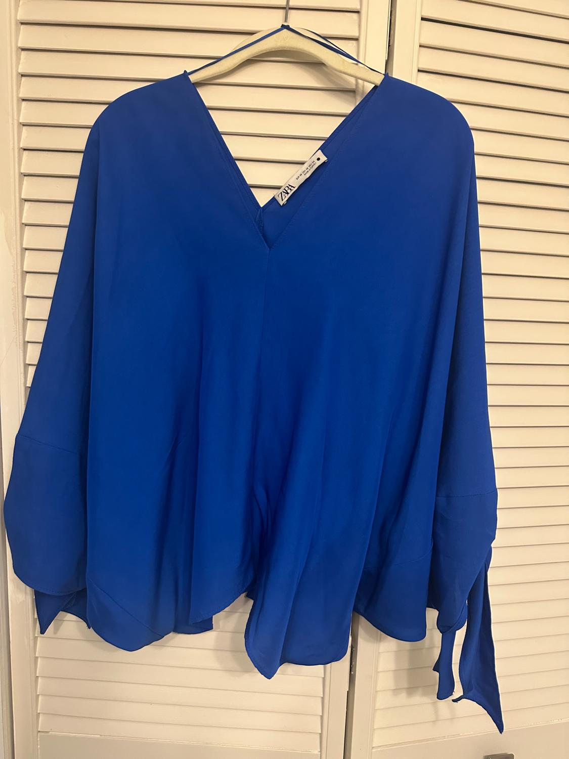 Flowing blue blouse with a V-neck and wide sleeves hanging on a hanger over white slatted doors. ZARA label visible.