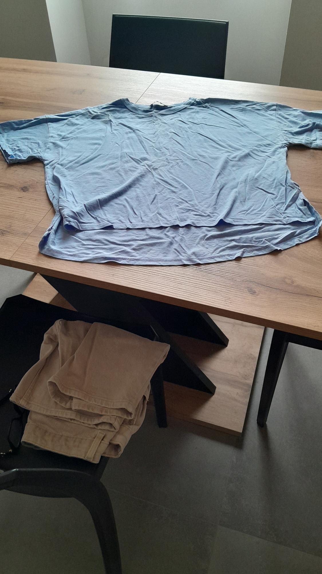Light blue wrinkled shirt on a wooden table, with beige trousers folded on a nearby chair. Indoor setting.