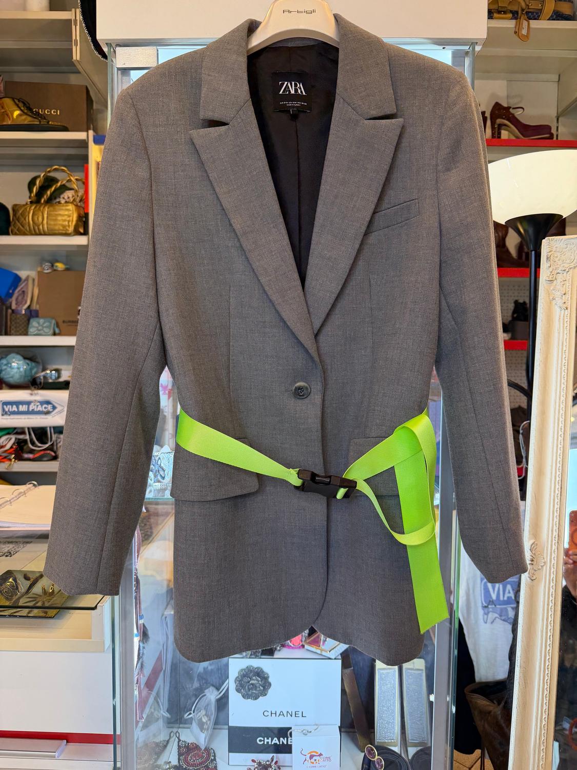 Zara grey jacket with a neon green belt, hanging on a hanger. Shop background with luxury accessories and boxes.