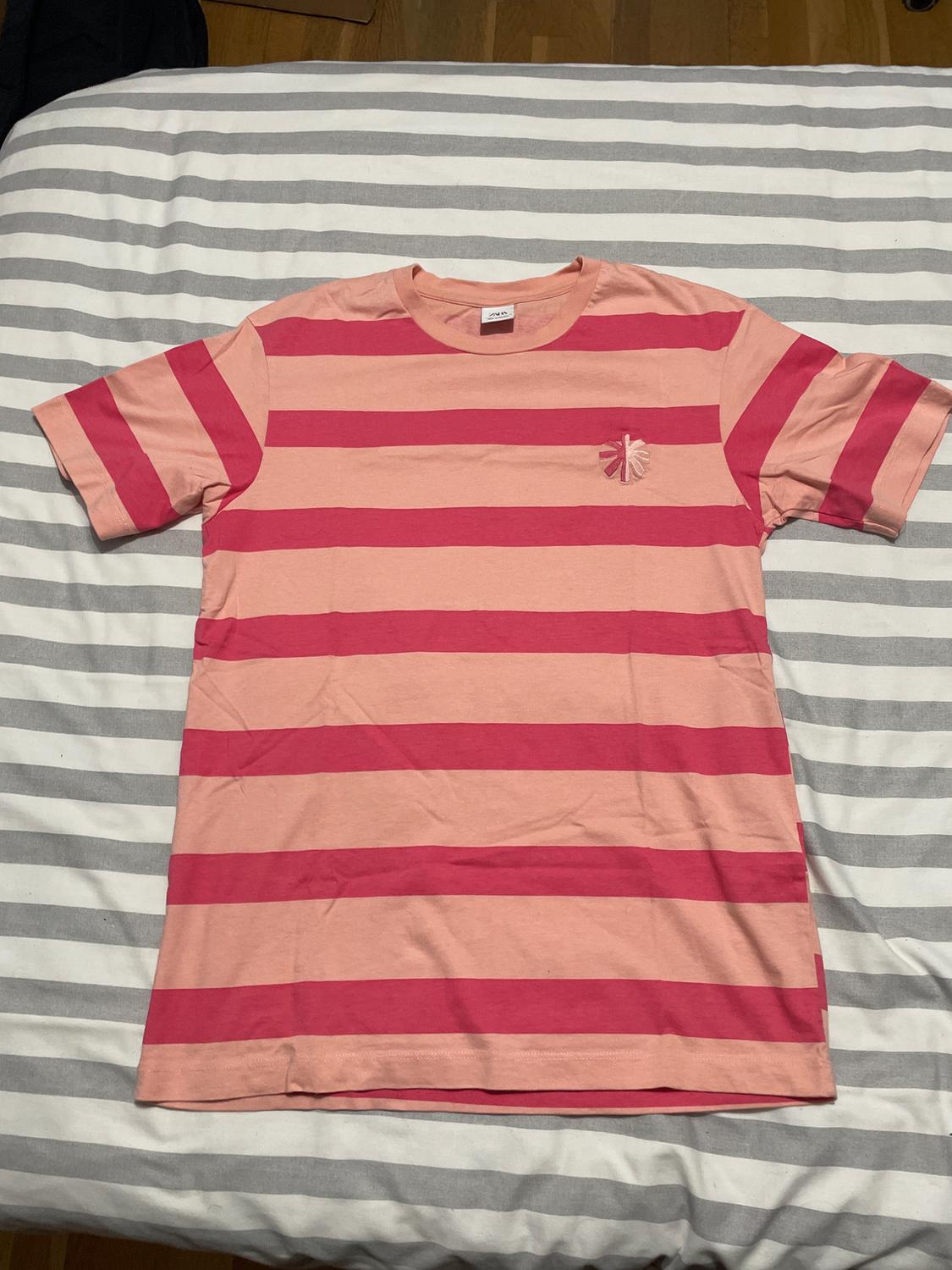 Pink and red horizontal striped t-shirt on a bed with grey and white striped sheets. Small logo on the chest.