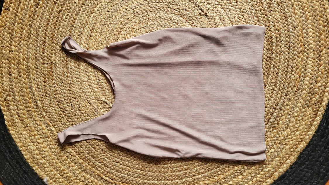Light beige sleeveless top on a braided jute rug. The garment is laid out flat and viewed from above.