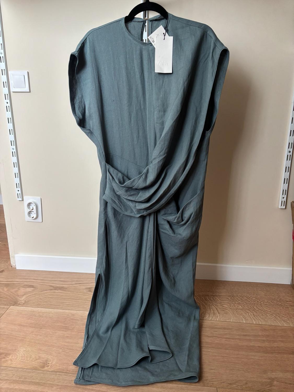 Greyish-green sleeveless dress with draping, side slit and ZARA label, on a black hanger against a beige wall and wooden floor.