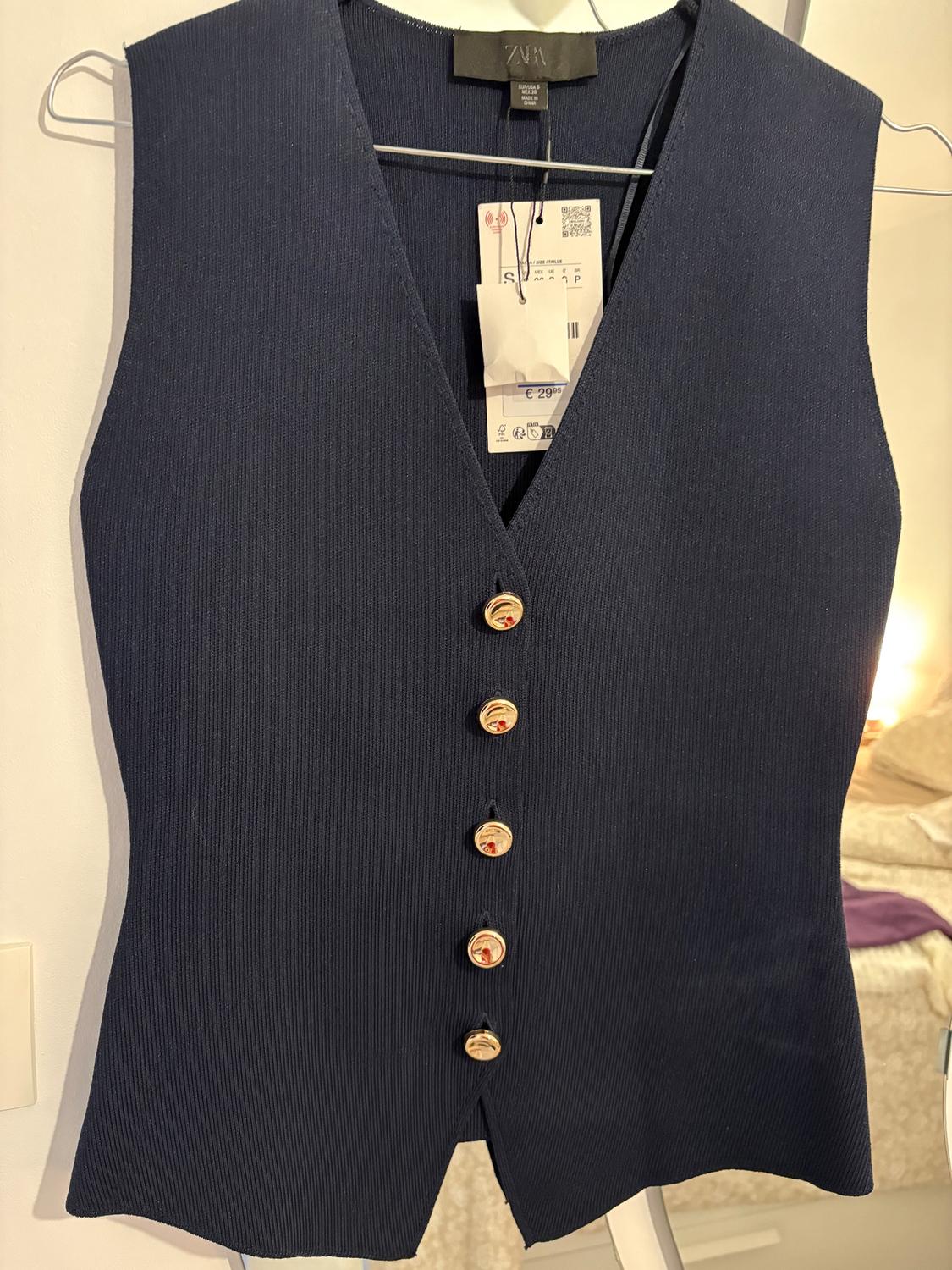 Dark blue waistcoat with gold buttons hanging on a hanger. Visible tag with price of €29.95. Room background.