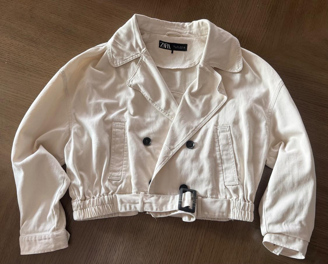 Cream-coloured short jacket on a wooden surface. Notable features include its dark buttons, elasticated hem with belt, and a label with the text ZARA.
