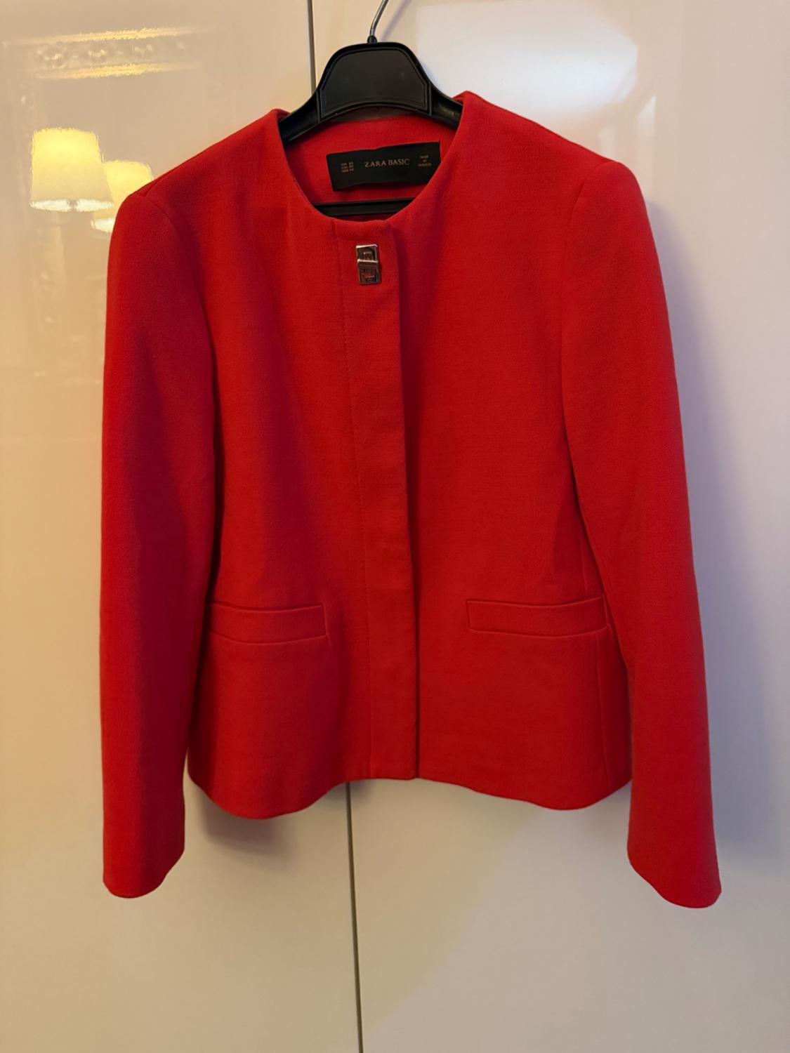 Red women's jacket hanging on a black hanger. Visible label: "ZARA BASIC". Background with lamp reflection.