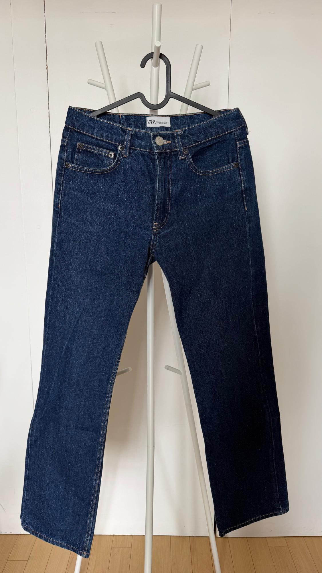 Dark blue jeans hanging on a black hanger on a white clothes rail. White wall background and wooden floor. ZARA label.
