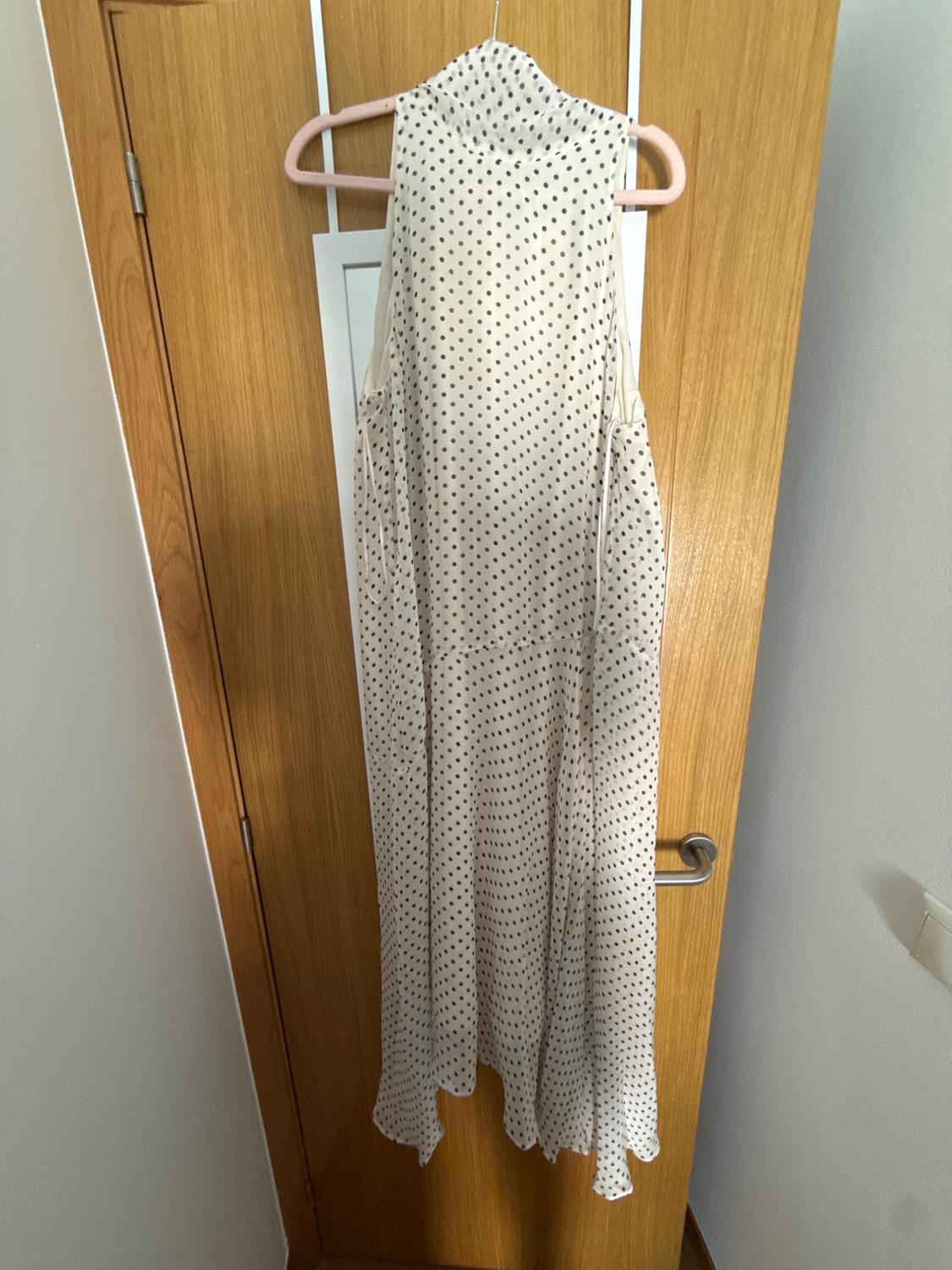 White sleeveless maxi dress with black polka dots, hanging on a pink hanger against a light wood door in a relaxed setting.