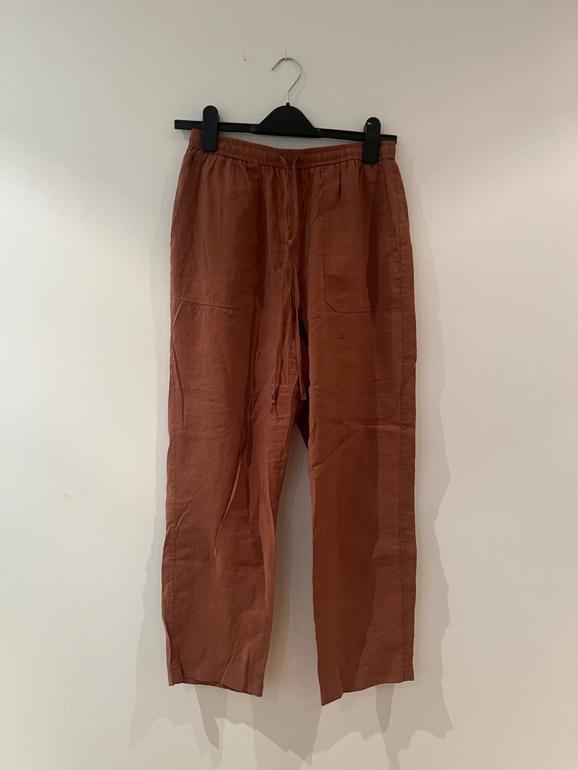 Brown trousers hanging on a black hanger against a white wall. Lightweight fabric with an elasticated waist and front pockets.