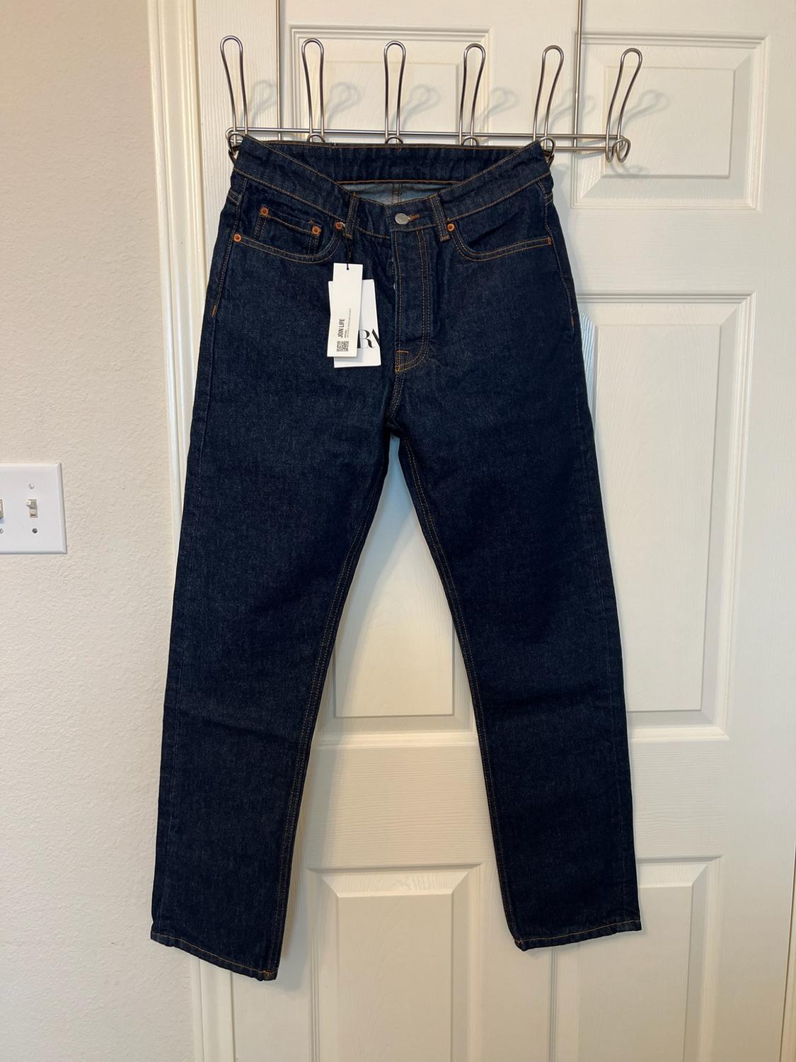 Dark blue jeans hanging from a hanger on a white door. They have a label bearing the text JOIN LIFE.