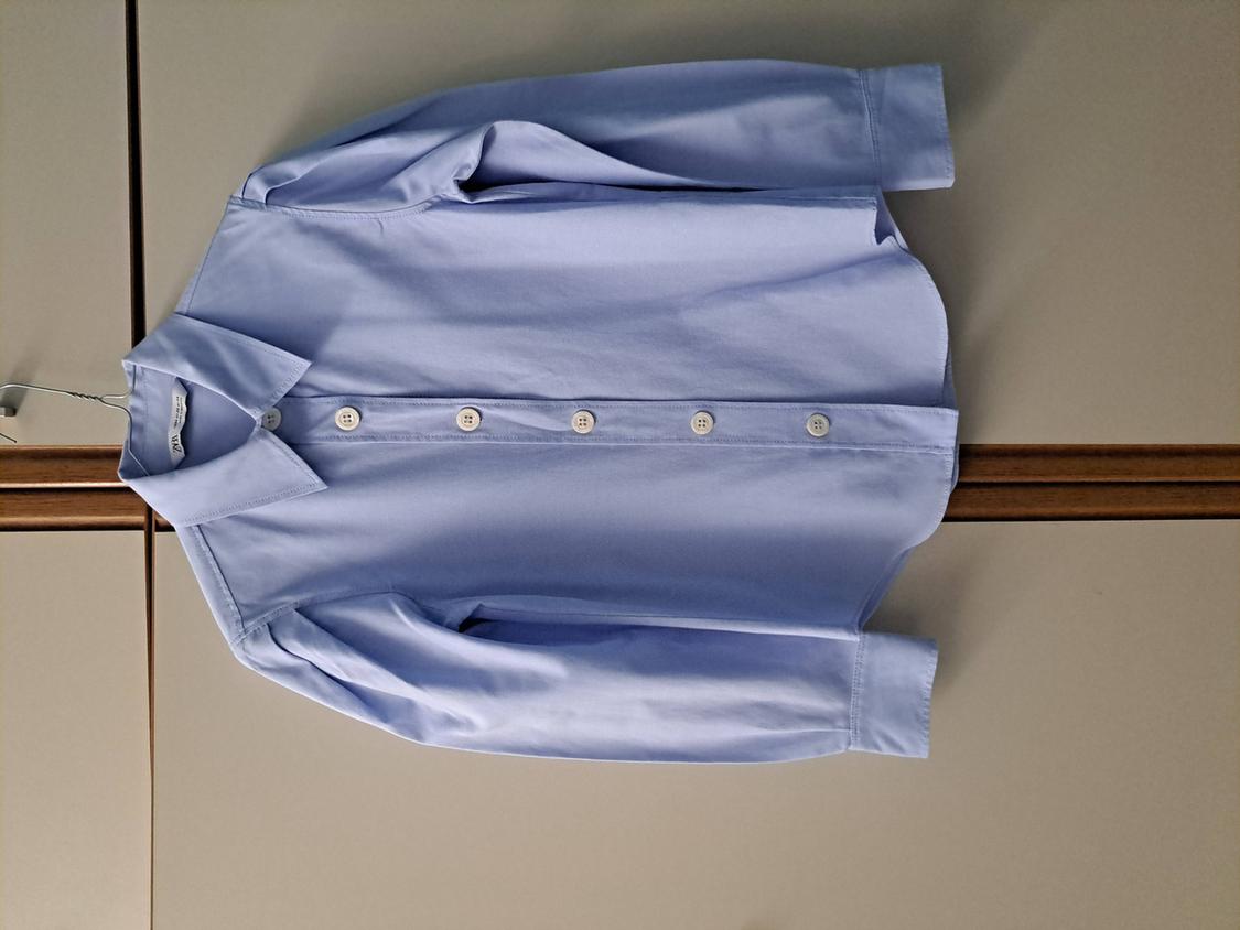 Light blue, long-sleeved shirt hanging on a hanger against a grey background. White buttons visible on the front.
