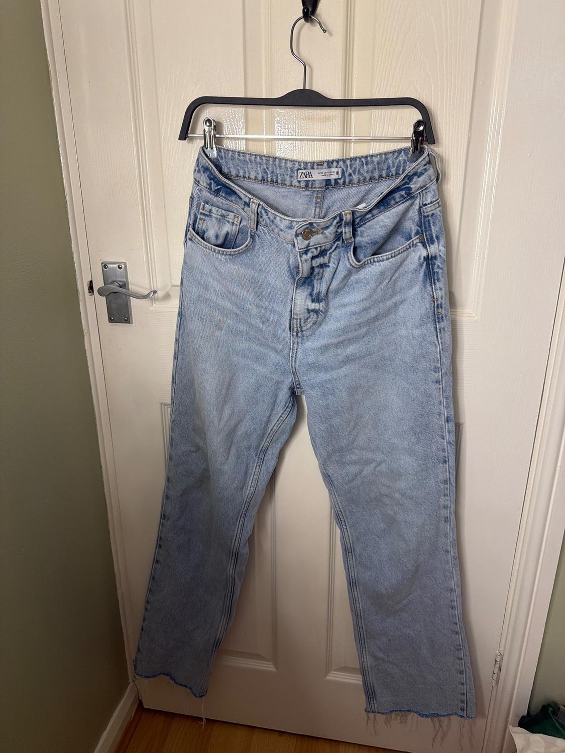 Light blue jeans hanging on a black hanger in front of a white door. They have a straight-cut design and frayed edges.