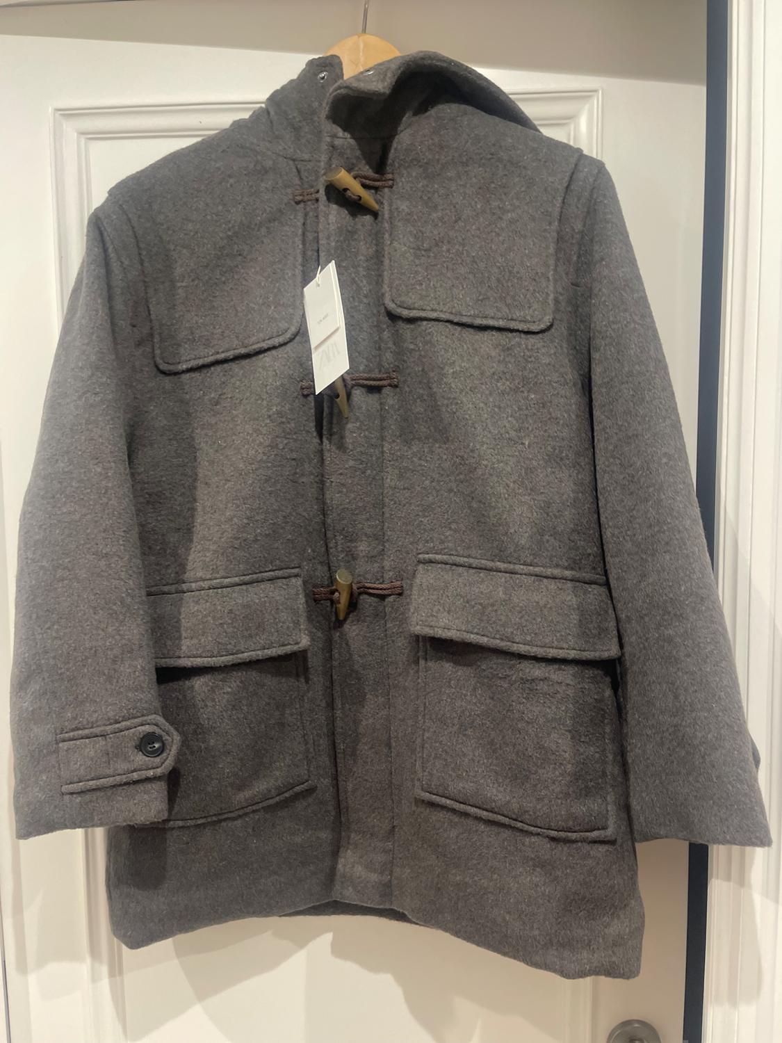 Grey duffle coat with a hood, hanging on a hanger over a white door. Visible label on the chest.