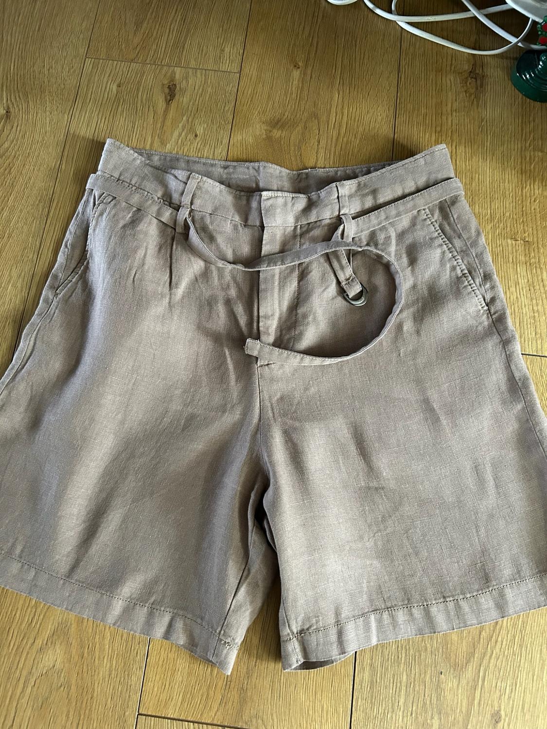 Beige tailored shorts with pleats, a matching belt and buckle, laid out on a light wood floor. Casual and relaxed style.