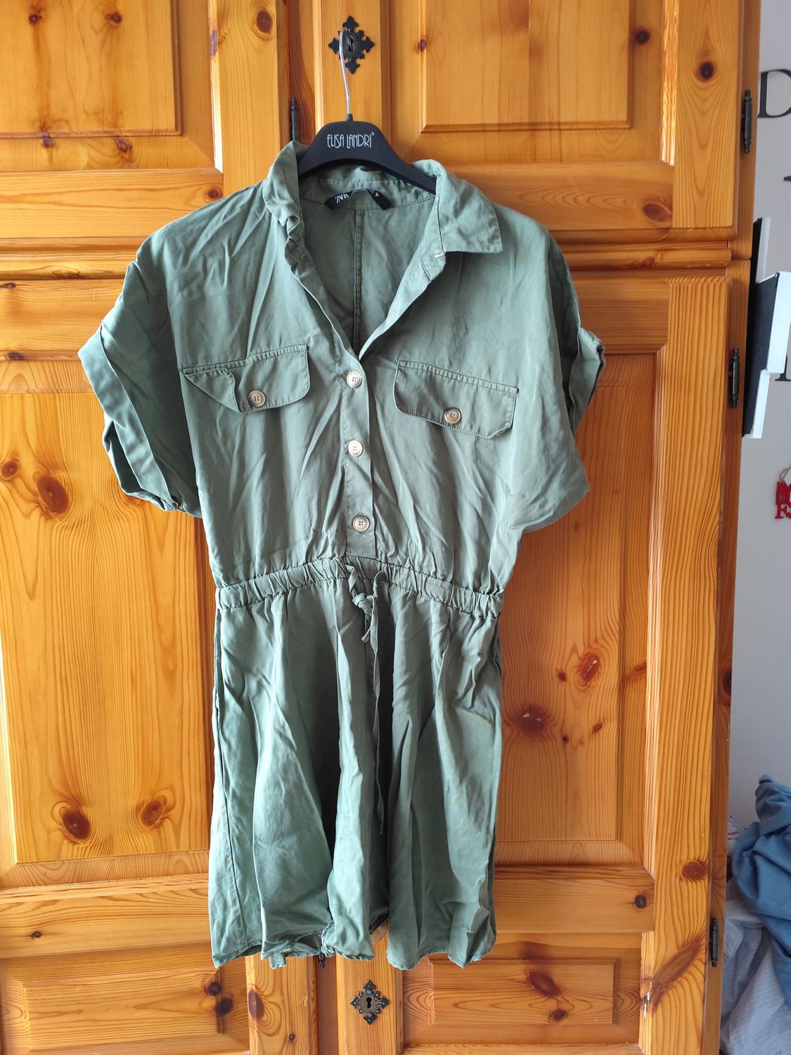 Olive green jumpsuit with buttons and chest pockets, hanging on a black hanger in front of a wooden wardrobe.