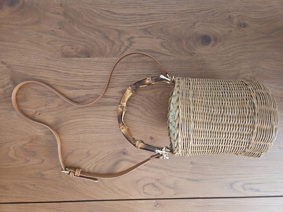 Cylindrical wicker bag with a brown leather handle and bamboo details on a light wooden surface.