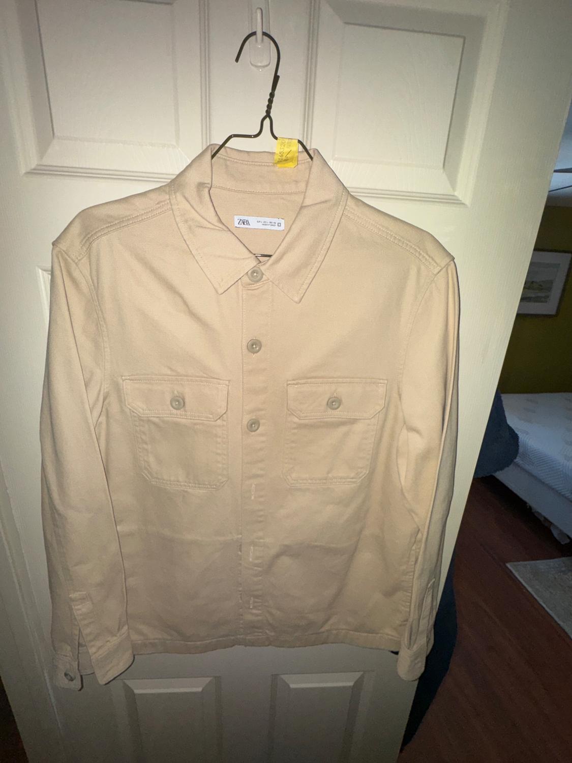 Beige long-sleeved shirt with two front pockets, hanging on a hanger in front of a white door. Visible tag: ZARA.