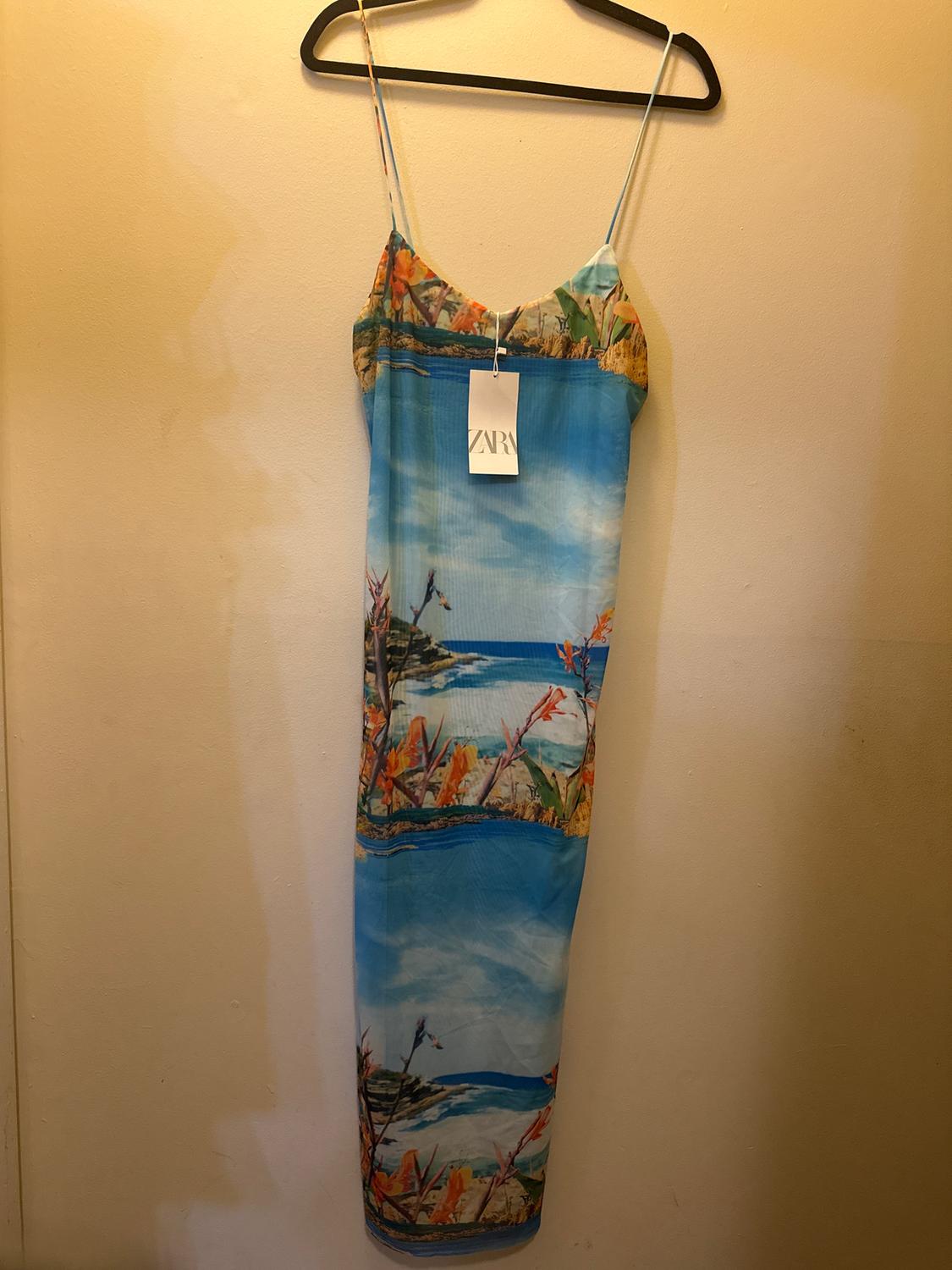 Long strap dress with a tropical landscape print in blue and orange tones, hanging on a hanger. Visible tag: ZARA.