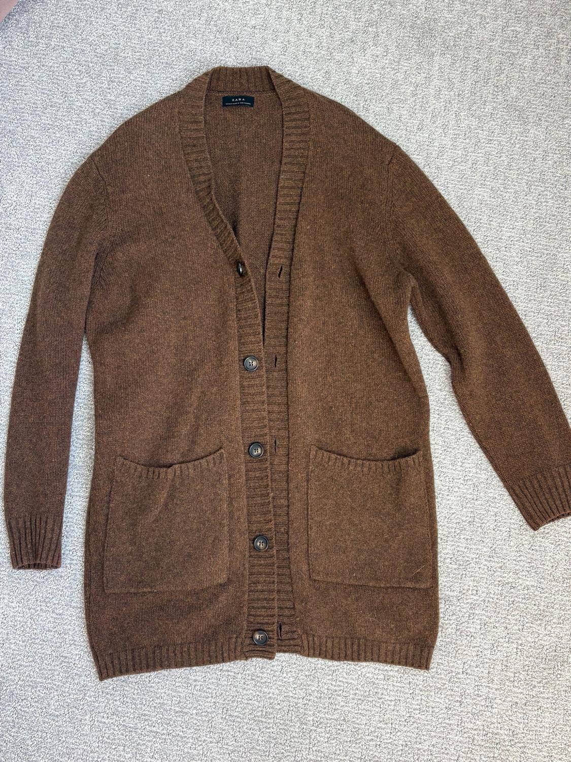 Brown knit cardigan with a pointed collar, buttons, pockets, and a ZARA tag, on a textured gray surface.