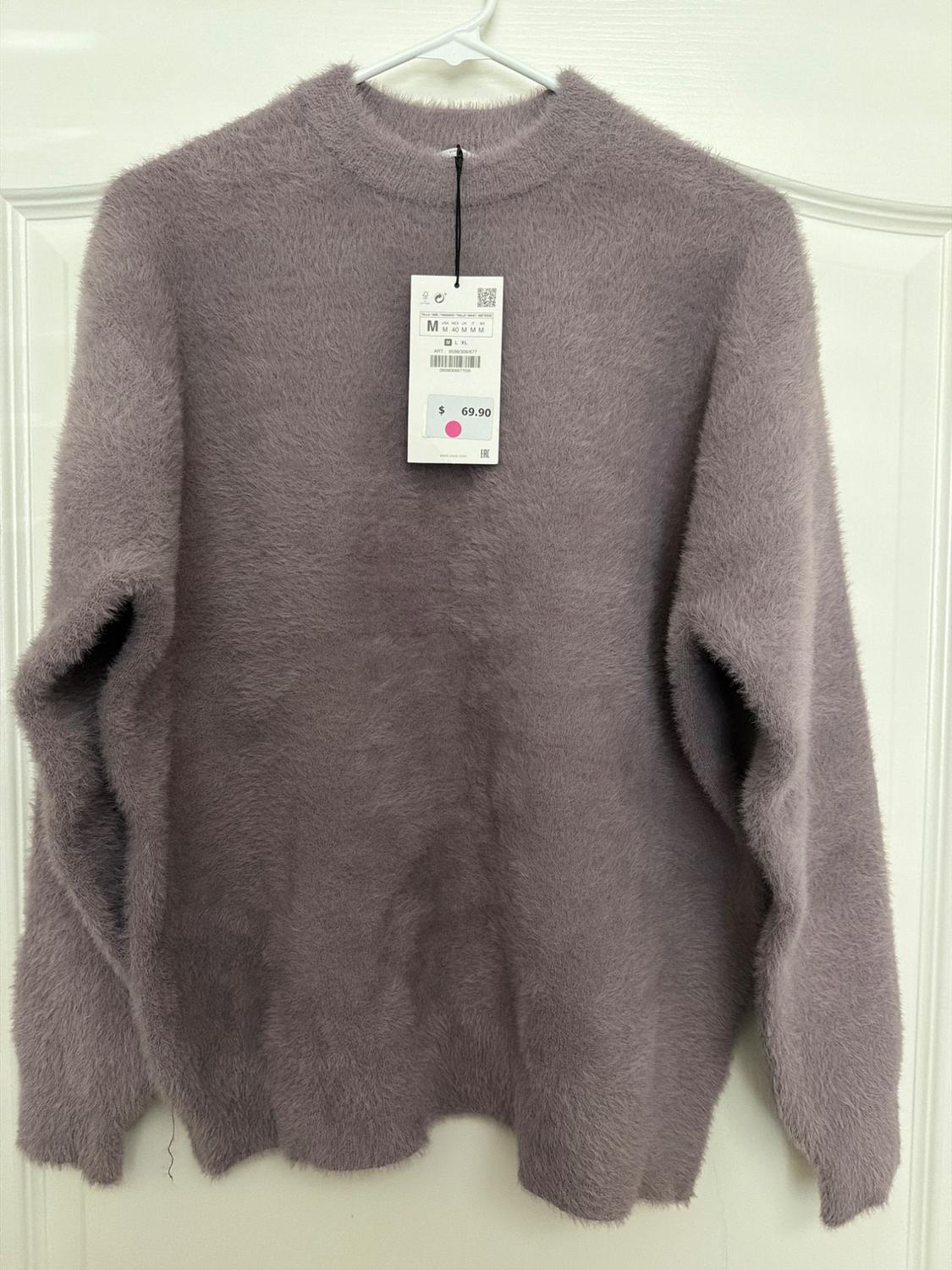 Gray fleece sweater hanging on a white hanger, with a price tag of $69.90 and size M, over a white door.