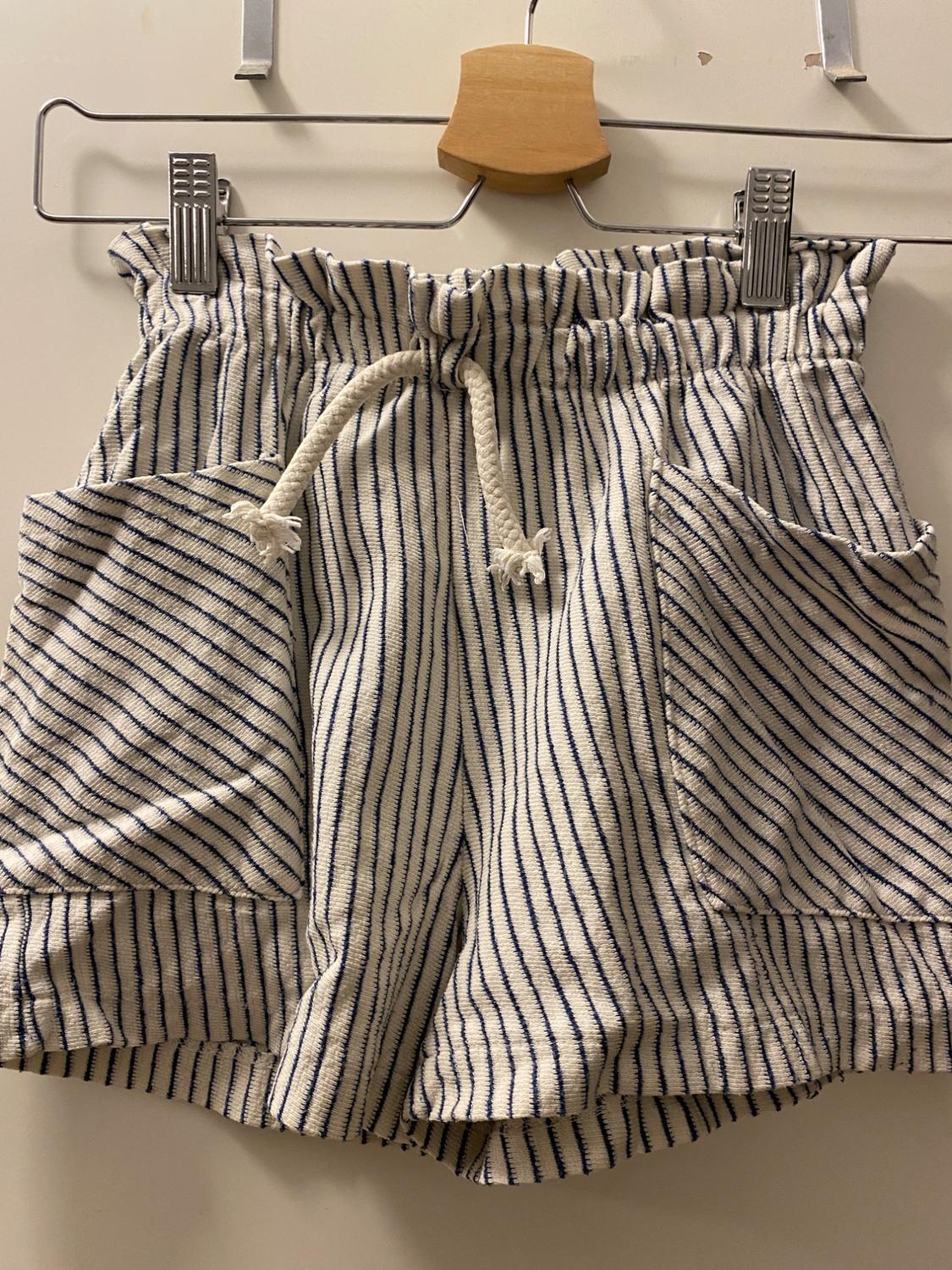 White and blue striped shorts with an elasticated waist and white drawstring, hung on a wooden hanger against a white background.