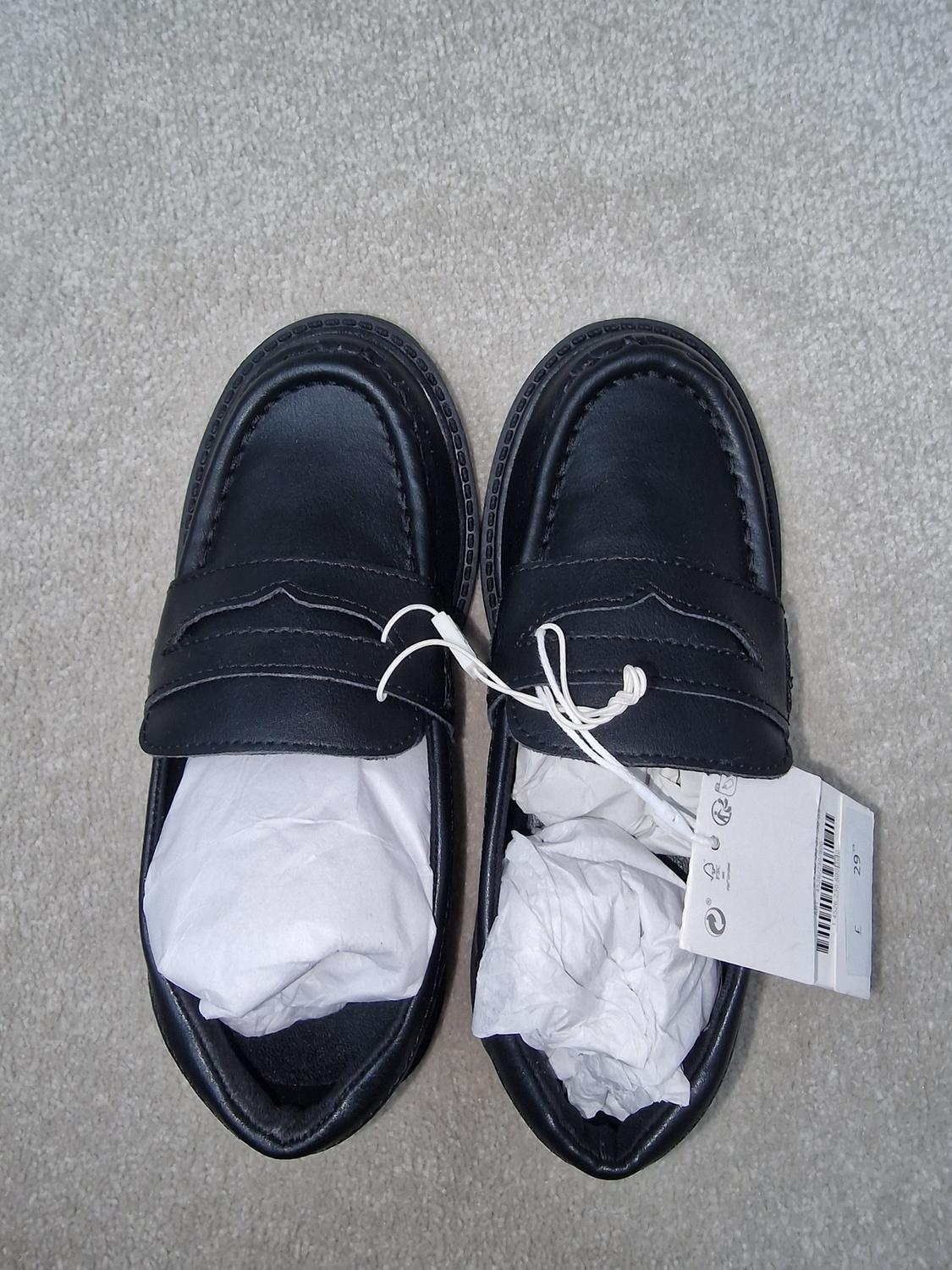 Black leather shoes with stuffing paper, seen from above on a grey carpet. White label with symbols and barcode.