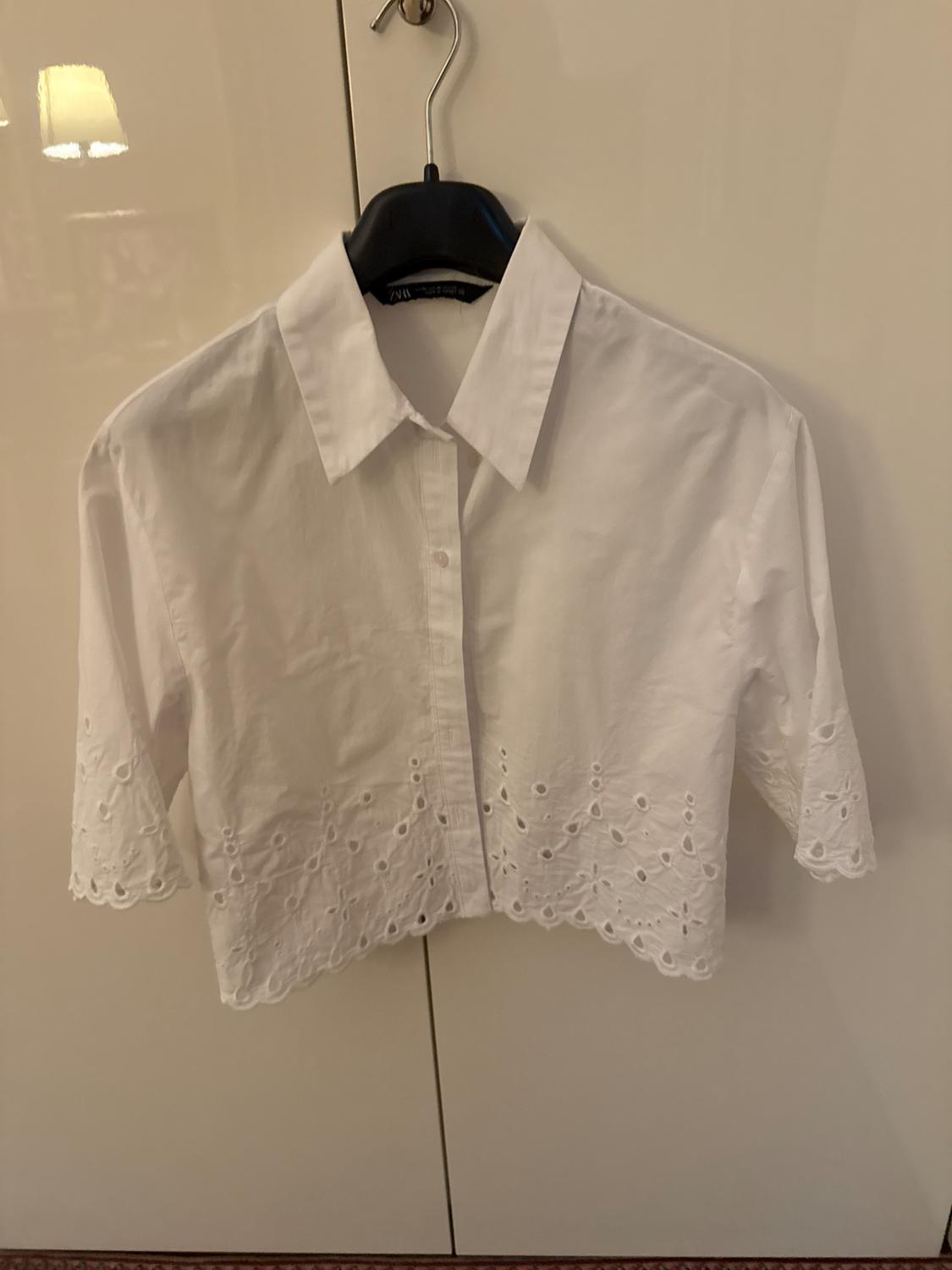 Short white three-quarter sleeve blouse with cutwork embroidery on the bottom edge, hanging on a black hanger in front of a light-coloured wardrobe.