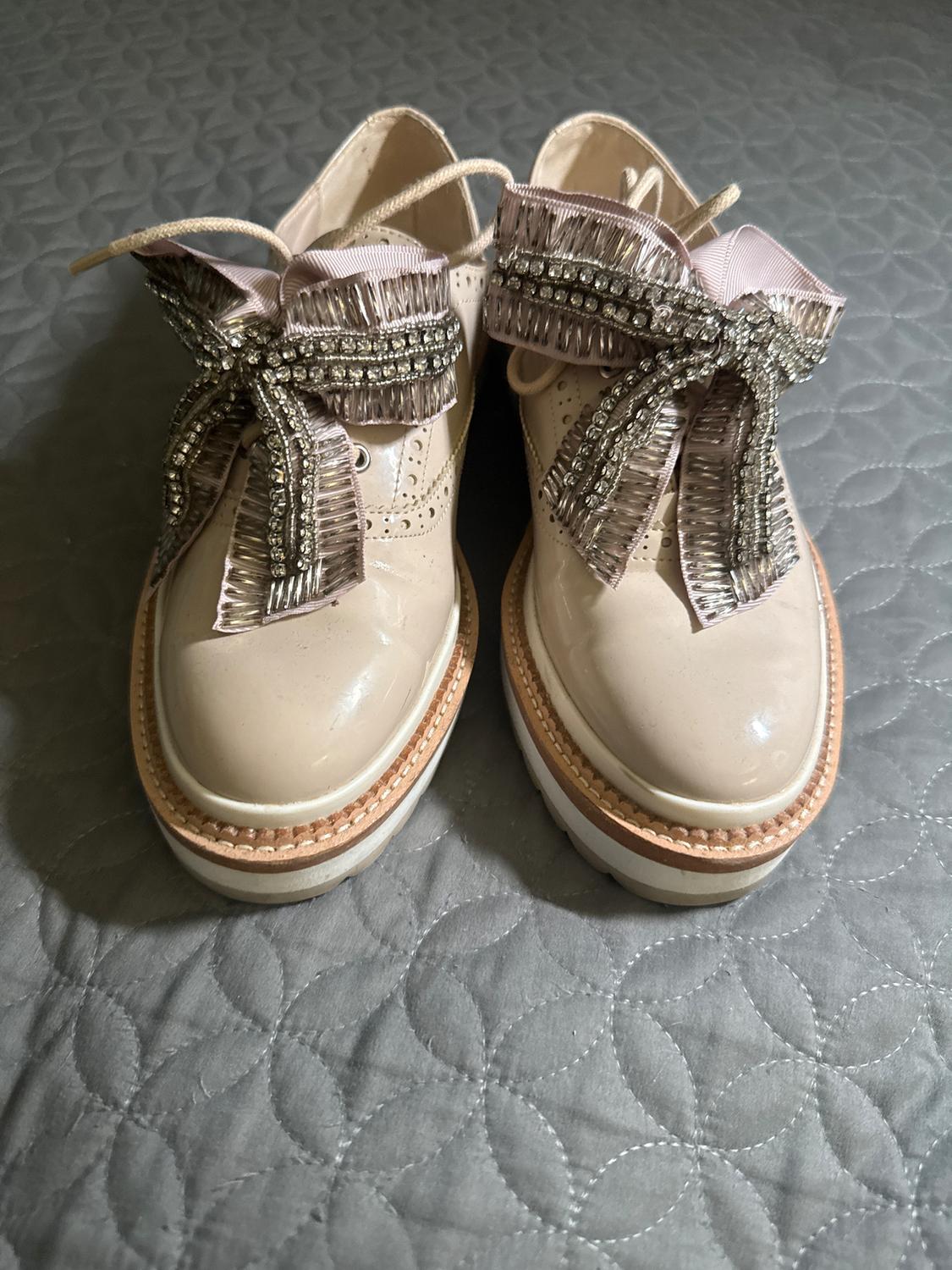 Beige shoes with decorative rhinestone bows and pleated ribbons on a gray comforter with an interlocking circle pattern.