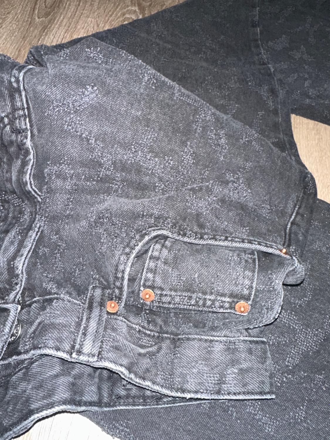 Dark textured denim pants with metallic button details on a light wooden floor.