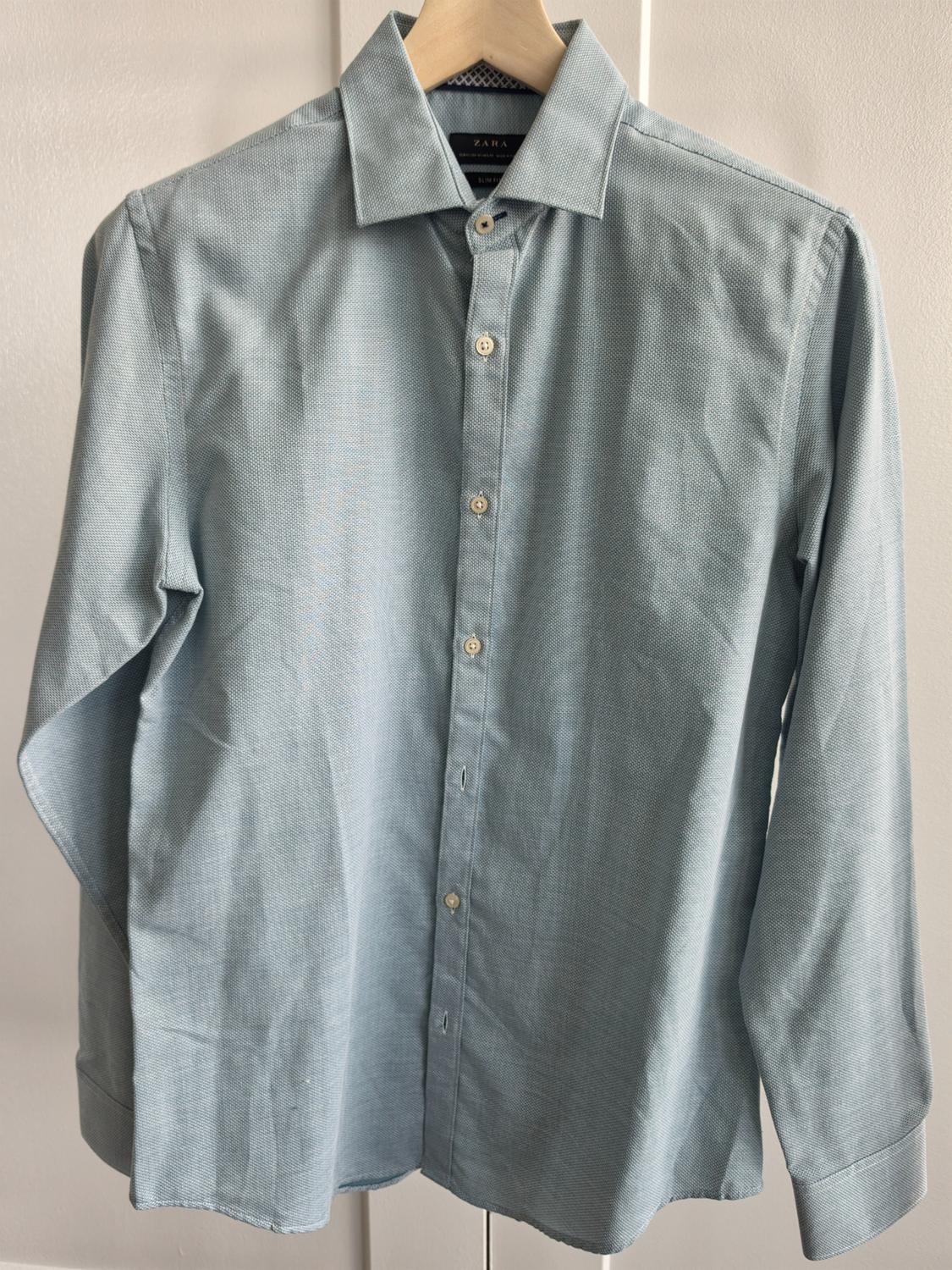 Light blue long-sleeved shirt hanging on a wooden hanger against a light background. The neck label reads ZARA SLIM FIT.