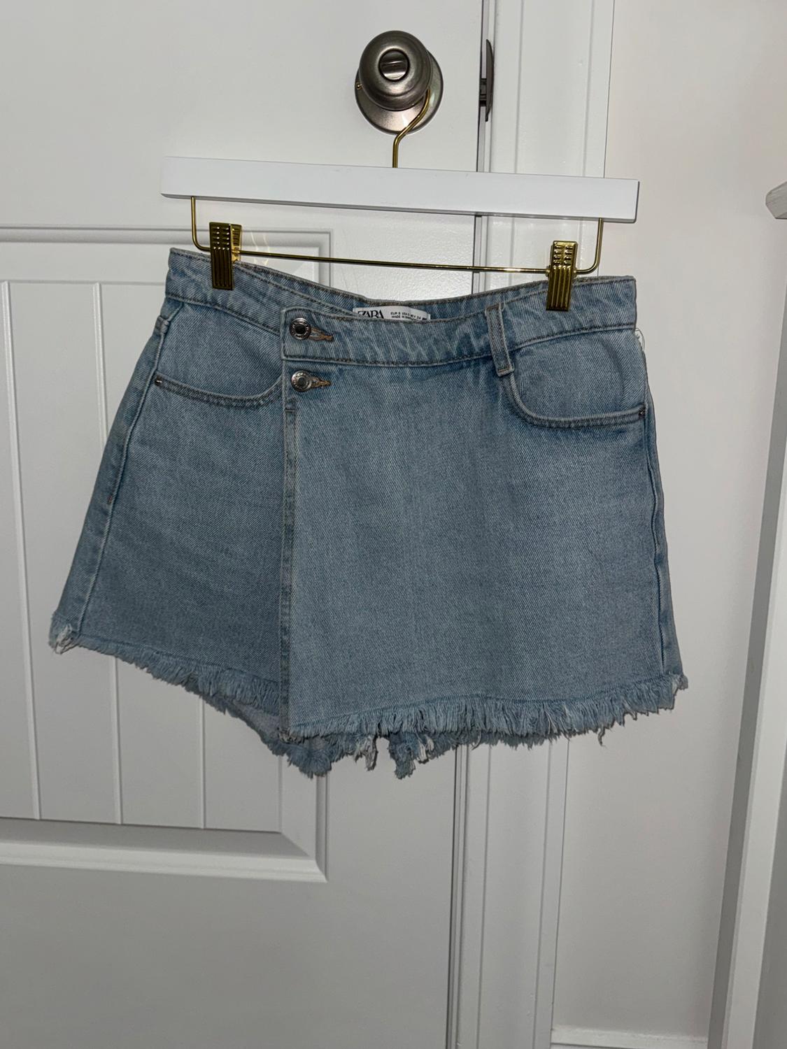 Light blue denim skirt with a frayed hem, hanging on a white hanger with gold clips in front of a white door.