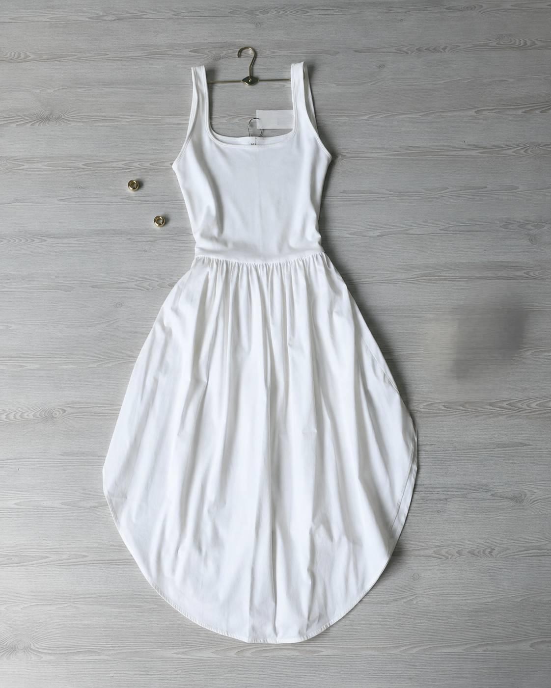 White, sleeveless, form-fitting dress with an asymmetrical skirt, presented on a grey wooden background with a hanger and gold accents.