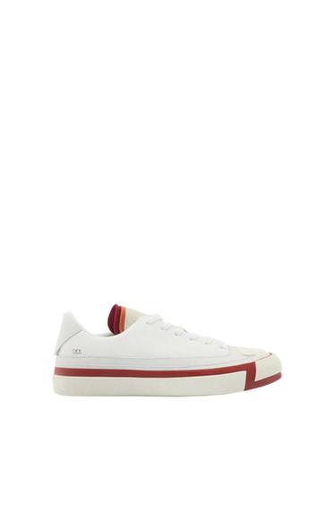 White side-profile trainer on a white background. Features laces, a red stripe on the sole, and warm details on the tongue.