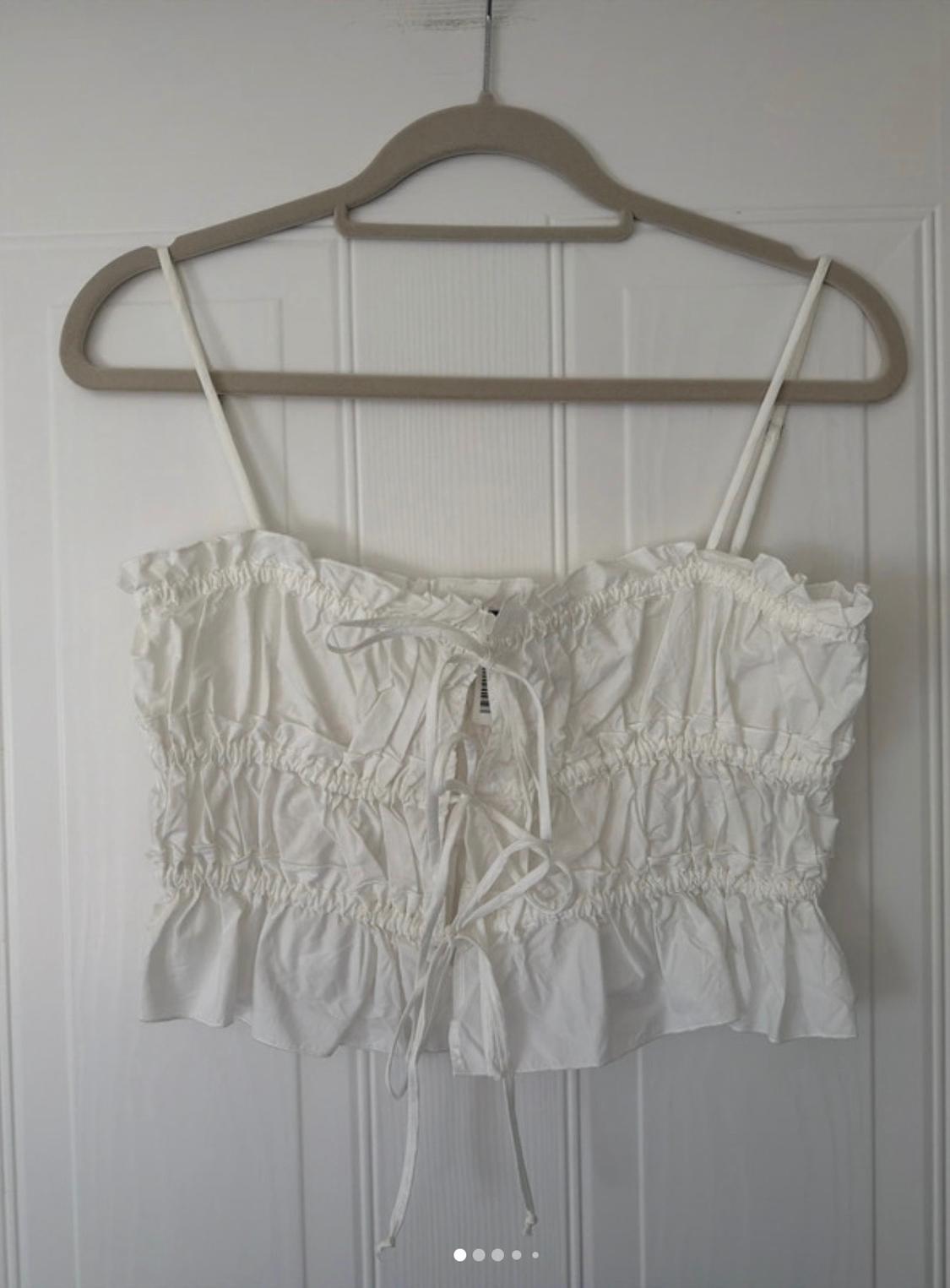 White strappy crop top with ruching and front ties, hanging on a hanger on a white door.